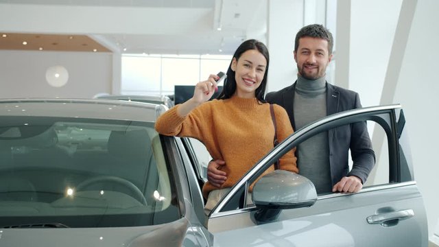 Portrait of happy young couple smiling standing beside expensive car in showroom holding key fob looking at camera. People, business and family concept.