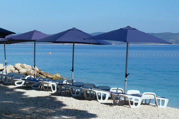 Fototapeta na wymiar Blue umbrellas and chaise for relax and comfort on sea beach. Happy summer vacations and tourism concept. Paid service on waterscape beaches.