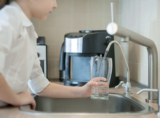 Little girl open a water tap with her hand holding a transparent glass. Kitchen faucet. Glass of clean water. Filling cup beverage. Pouring fresh drink. Hydration. Healthcare. Healthy lifestyle