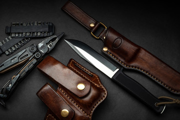 Hunting knife and multitool on a dark background. Brown leather cases near the knife and multitool. Postcard concept for men.