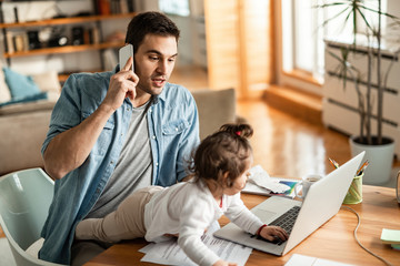 Young working father with small daughter talking on mobile phone at home.