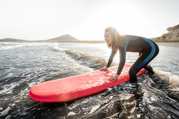 Young surfer in wetsuit getting on surfboard, catching water flow near the beach during a sunset. Water sports and active lifestyle concept