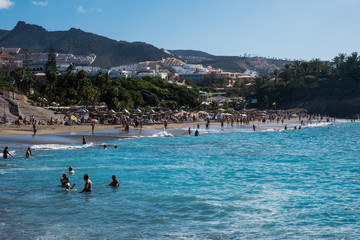COSTA ADEJE, TENERIFE, SPAIN - NOVEMBER 2019: People lay on the sand and swim in the ocean at tropical beach.