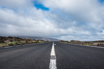 Fototapeta premium Endless road to Timanfaya National park in Lanzarote, Canary Ispands, Spain, Europe, Africa. Volcanic, black sand, harsh, tough, inhospitable, dry, sub-tropical, desert landscape.