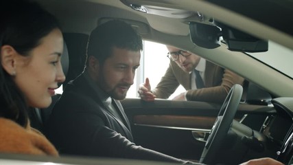 Smiling young couple inside new car talking to sales manager in dealership choosing new automobile enjoying high quality of model. People and vehicles concept. - Powered by Adobe