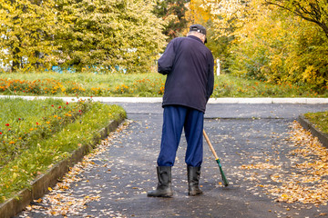 The janitor sweeps the fallen yellow leaves on the road. Cleaning leaves from the gutter with a...