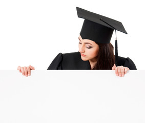 Female graduate student peeking from behind a blank panel, isolated on white background. Beautiful graduate teen girl student in mantle showing blank placard board.
