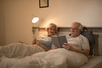 Mature man reading a book while resting with his wife in bedroom at night.