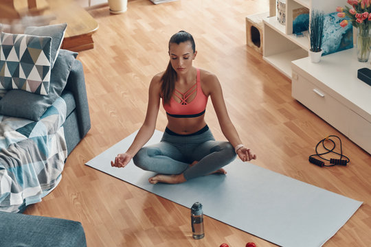 Top View Of Beautiful Young Woman In Sports Clothing Meditating While Sitting In Lotus Position At Home