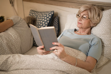 Senior woman relaxing while reading a book in bedroom.