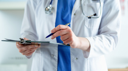 Doctor with a stethoscope, holding a notebook in his hand. Close-up of a female doctor filling up medical form at clipboard while standing straight in hospital