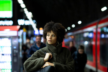 Stylish portrait of a beautiful African-American woman with black curly hair in a fur coat. Woman standing on urban city subway metro platform at night. Fashion and lifestyle