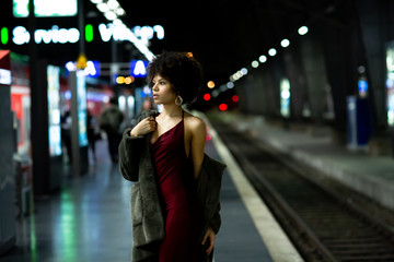 Young stylish African-American model woman in red dress and a fur coat posing on the subway...