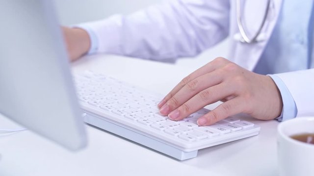 A Young female woman doctor is working at her office with computer and typing electronic medical record system, white table background, close up, crop view