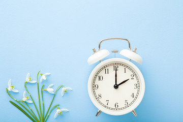 White alarm clock with fresh, beautiful snowdrops on light blue table background. Pastel color. First messengers of spring. Time concept. Closeup. Empty place for text. Top down view.
