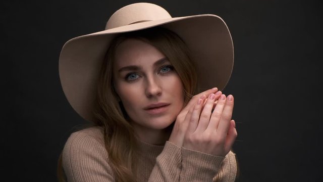A young beautiful elegant woman in a beige sweater and fedora with fields poses against a black background. A close-up of the face. Romantic and sensual lady.