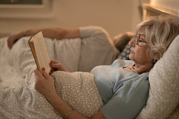 Senior woman reading a book while resting in bed in the evening.