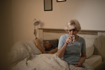 Senior woman drinking water while sitting on the bed in the evening.
