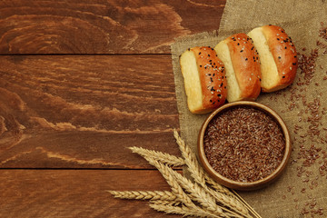 three small rolls of bread sprinkled with sesame seeds lie on a linen napkin, and next to it is a bunch of wheat ears and a bowl of flax seeds on a brown wooden background view from the top