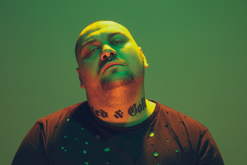 Portrait of a hipster guy with colorful neon light on green studio background. Male model with calm and serious mood. Facial expression, cyberpunk, millenials lifestyle and look like. Fashion.