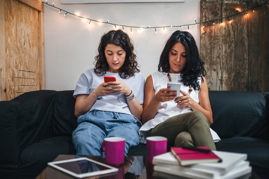Two young women on the sofa at home look their smart phones - Millennials have fun browsing on social networks