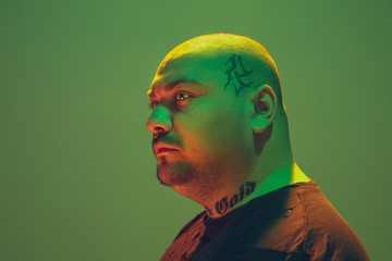 Portrait of a hipster guy with colorful neon light on green studio background. Male model with calm and serious mood. Facial expression, cyberpunk, millenials lifestyle and look like. Fashion.