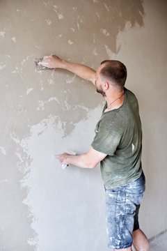 Male worker plastering wall with putty-knife, close up. Fixing wall surface and preparation for painting. Young man with a beard in a T-shirt and shorts smeared with paint