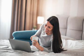 Girl lying on the bed drinking coffee and using laptop.