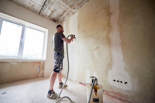 Man painting a grey wall with spray gun, renovating of new house. Open container with a paint. Spray paint work. Male is dressed in paint-smeared clothing. Small room with window and daylight