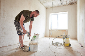 Worker mixing dry mortar with handheld mixer in bucket at repairable room. Man in smeared clothing kneads a solution against the background of a window in daylight