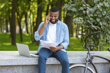 Fototapeta premium Black man sitting in park talking on cellphone and checking documents