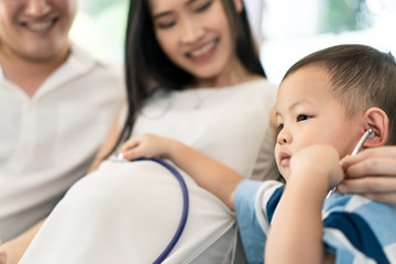 Obraz premium Asian family sitting together at home with smile. Young child listening baby heart pulse on mother tummy by stethoscope with father. Kid feeling excited with future coming newborn. Family relationship
