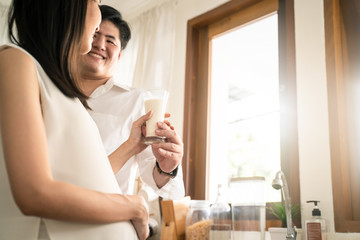 Asian young couple standing in kitchen. Man take care pregnant woman by giving milk. Mother drink calcium milk for little baby health in tummy. Love and health care pregnancy girl for future newborn.