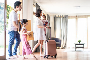 Asian big family moving packing boxes and cloth baggage to new house. Mother holding little cute baby girl with suitcase while dad and bigger sister carrying box walking into house with happy, smile.