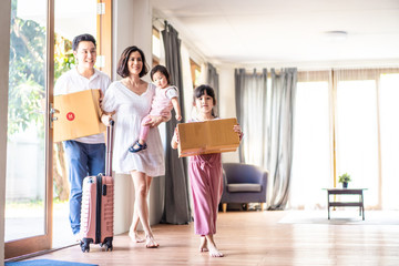 Asian big family moving packing boxes and cloth baggage to new house. Mother holding little cute baby girl with suitcase while dad and bigger sister carrying box walking into house with happy, smile.