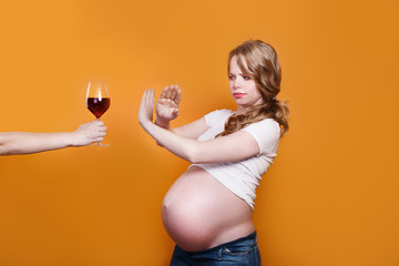 Pregnant woman refuses to drink wine, making stop gesture to glass on yellow background