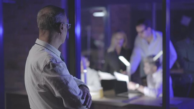 Mature businessman looking through glass wall at his colleagues