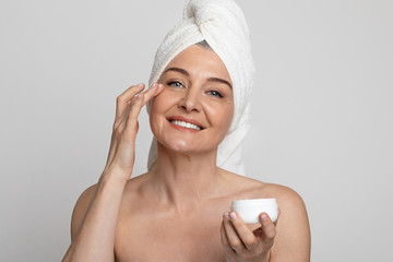 Happy mature woman applying anti-aging cream on face over light background