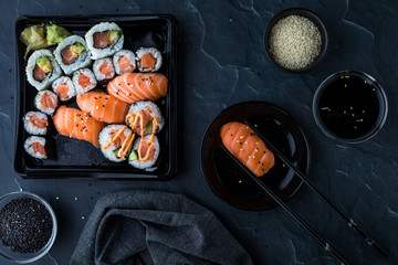 A top down view of assorted sushi against a dark background and surrounded by sesame seeds, soy sauce and chopsticks. © Carey