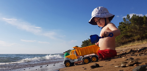 a little boy playing with toy truck and shovel on the beach on a hot summer day