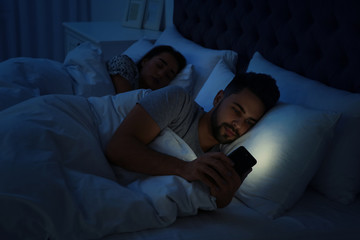 Fototapeta na wymiar Young man using smartphone while his girlfriend sleeping in bed at night