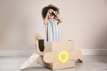 Cute African American child playing with cardboard plane and binoculars near beige wall