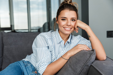 Image of happy caucasian woman smiling at camera while sitting on sofa