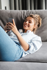 Image of smiling nice woman using cellphone while lying on sofa