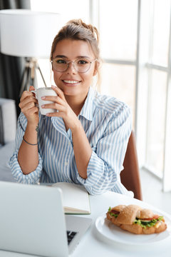 Image of joyful woman drinking coffee and using laptop while sitting