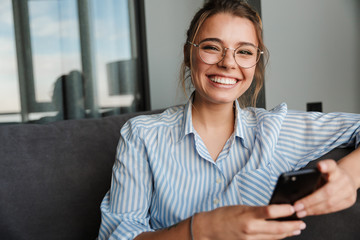 Fototapeta Image of happy young woman in eyeglasses smiling and using cellphone obraz