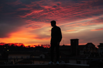 Fototapeta premium Silhouette of man standing and looking the vision over the cityscape background at sunset time. Orange, rose and blue clouds.