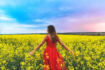 Fototapeta premium Young beautiful girl in a red dress close up in the middle of yellow field with radish flowers at sunset time