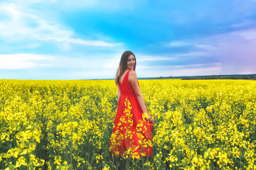 Naklejka na ściany i meble Young beautiful girl in a red dress close up in the middle of yellow field with radish flowers at sunset time