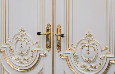 Close-up of white baroque doors with classic golden handles and ornaments.